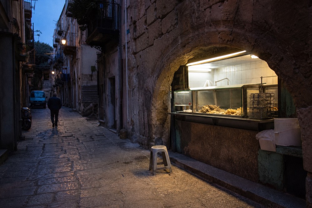 A quiet Palermo alley at dusk with a single lit friggitoria counter and a few stools