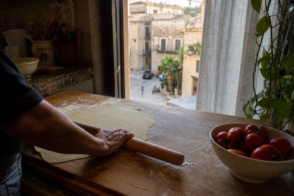 Fresh pasta dough and a rolling pin on a floured wooden board beside a bowl of tomatoes