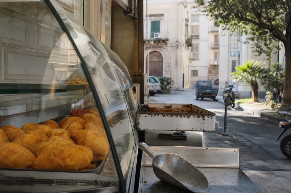 A Palermo bakery counter with rows of round fried snacks under glass
