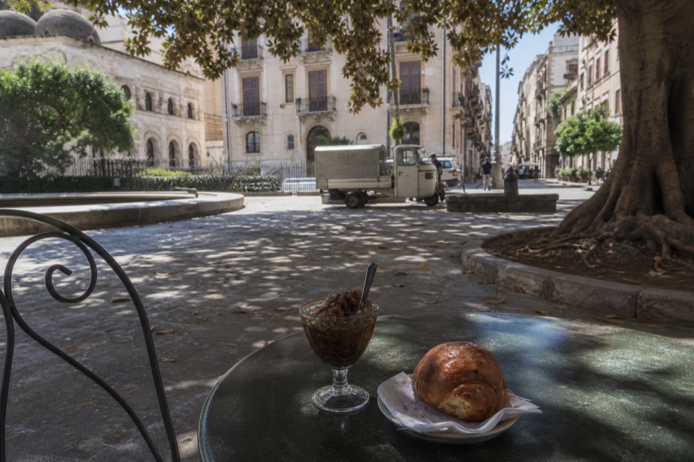 A glass of coffee granita with a sweet brioche resting on the saucer of a cafe table