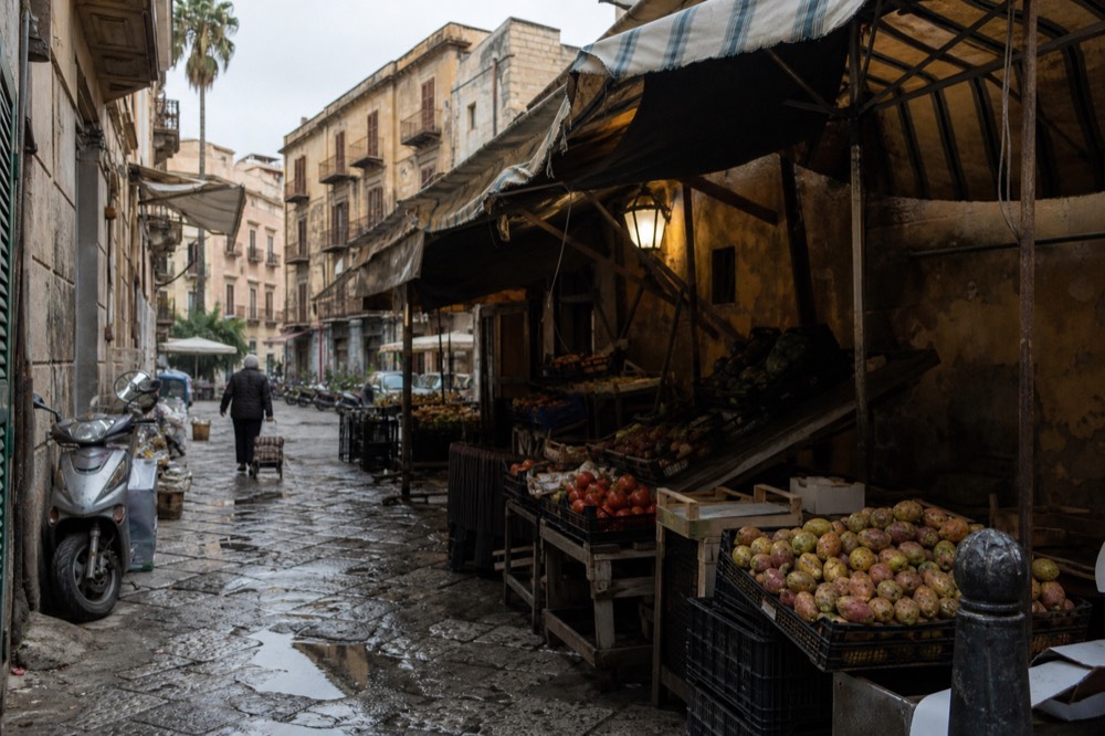 Market stalls with fruit and vegetables displayed under awnings in Ballarò