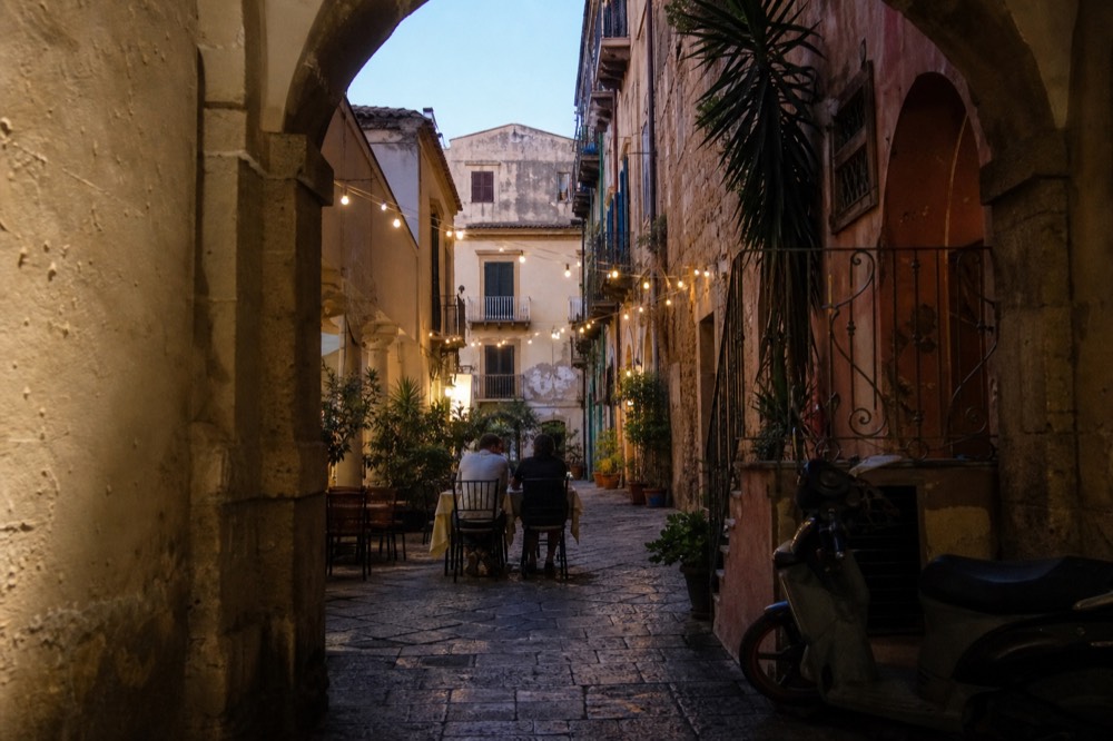 A narrow evening street in Palermo with cafe tables and lit old buildings