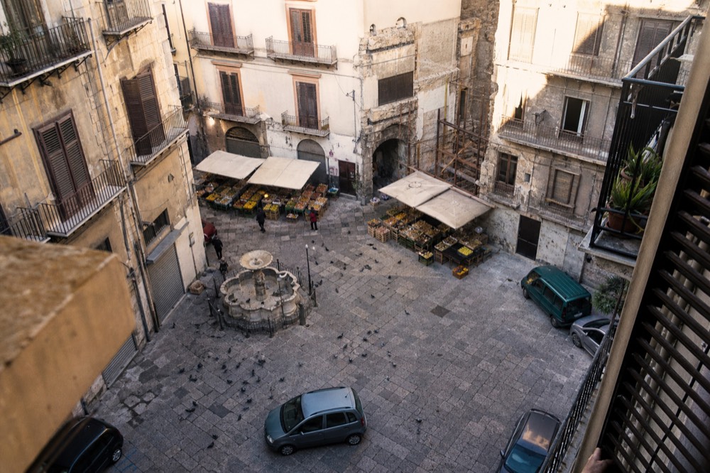 A sunlit Palermo market square with worn stone buildings and a few open stalls
