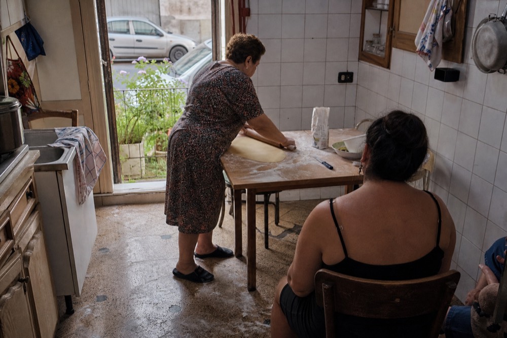 A home kitchen with an older woman rolling dough while guests watch from stools