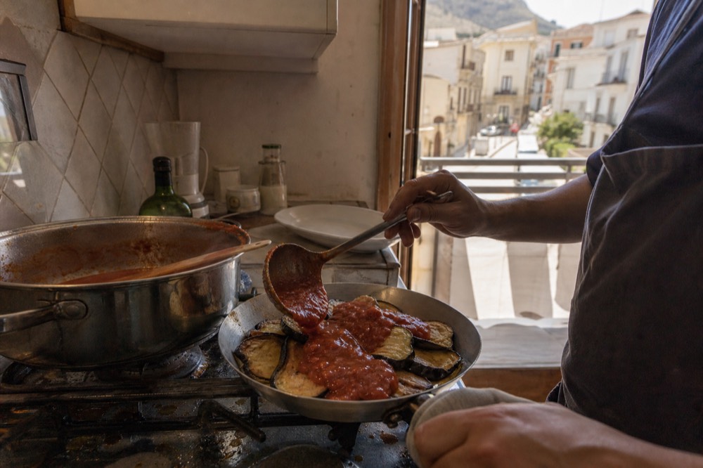 A cook ladling tomato sauce over a pan of fried aubergines