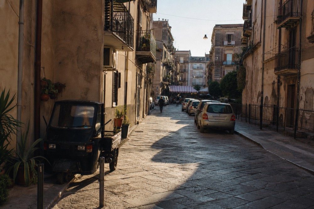 A sunny Palermo street with balconied buildings and a market awning in the distance