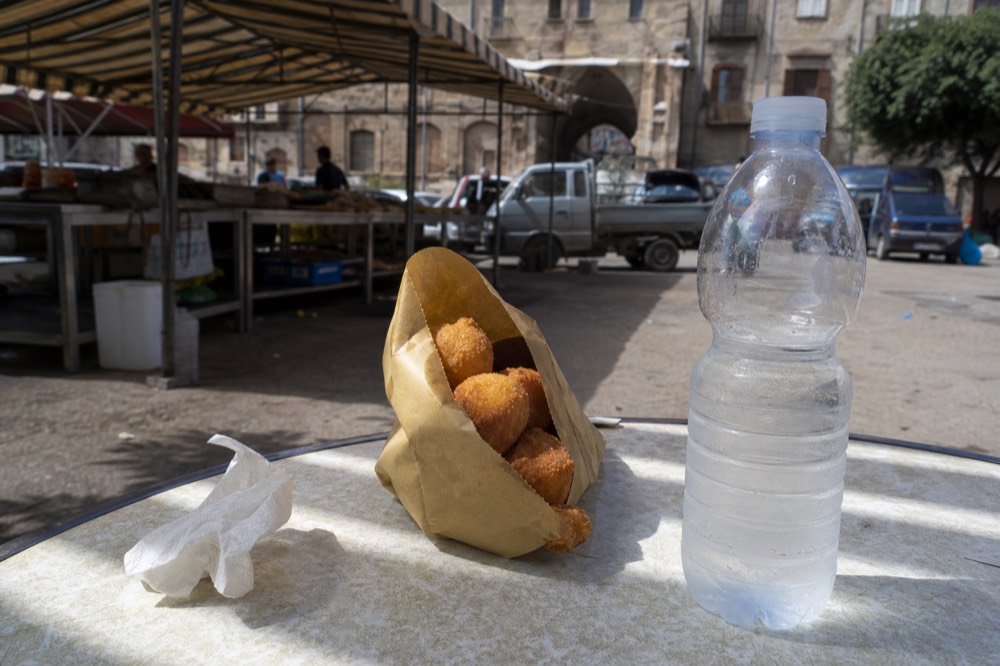 Paper cone of fried potato croquettes beside a bottle of water on a market table