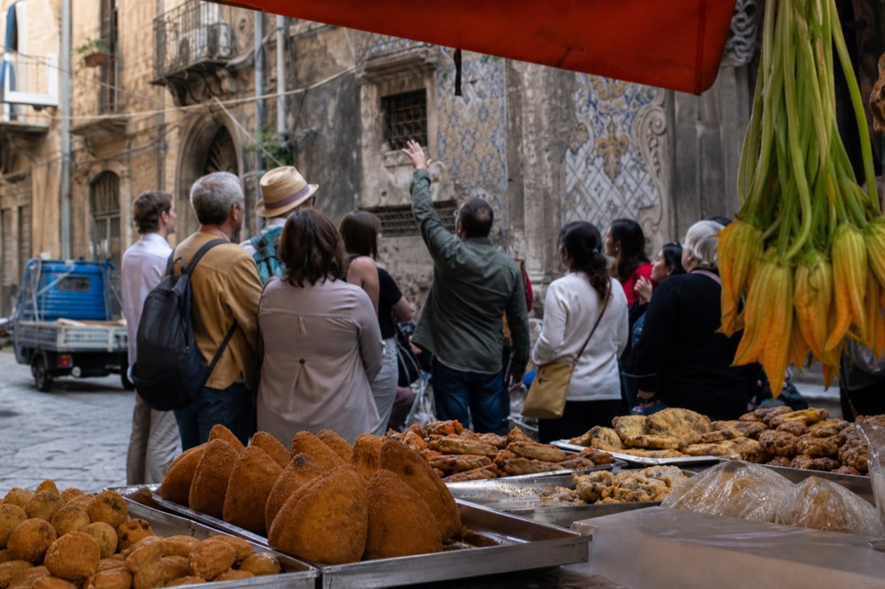 A small group standing at a market stall while a guide points at trays of food