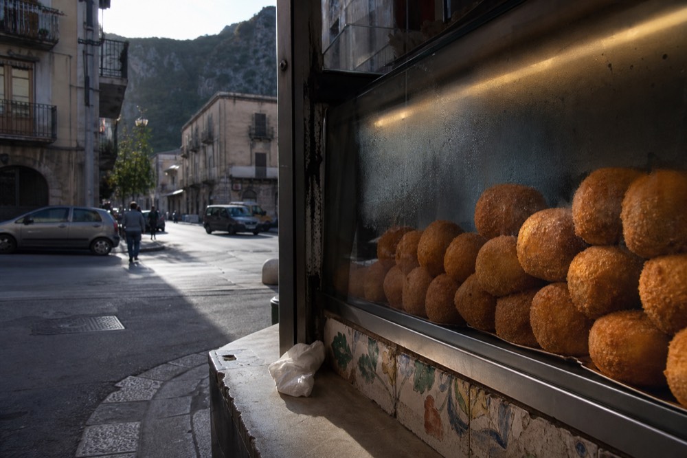 Round golden fried arancine stacked in a glass display case at a Palermo bakery