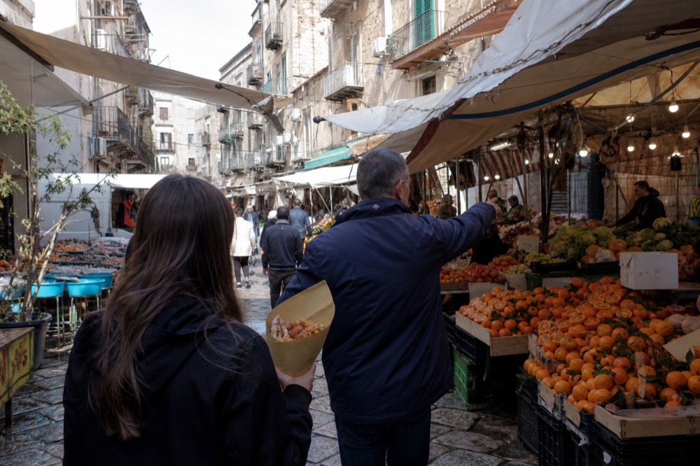 A small group of walkers sampling food from a market stall with a guide gesturing at the produce