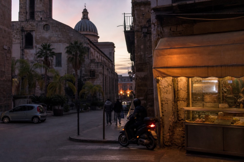 A Palermo street corner with a small food shop's awning and evening light on old stone buildings